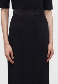 Personne portant une jupe noire taille haute et un haut noir, debout les bras détendus le long du corps sur un fond uni et clair.