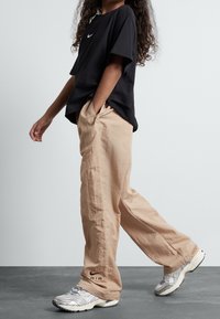 Černé tričko s krátkým rukávem a bílým logem, béžové volné kalhoty a šedé tenisky s texturovanými detaily a značkováním na boku.