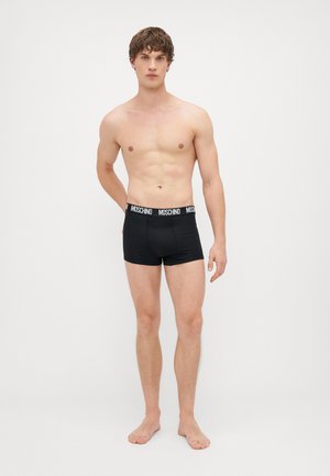 Fekete boxer alsónadrág rugalmas anyagból, puha, elasztikus derékpánttal, fehér "Moschino" márkázással, modell mezítláb áll.