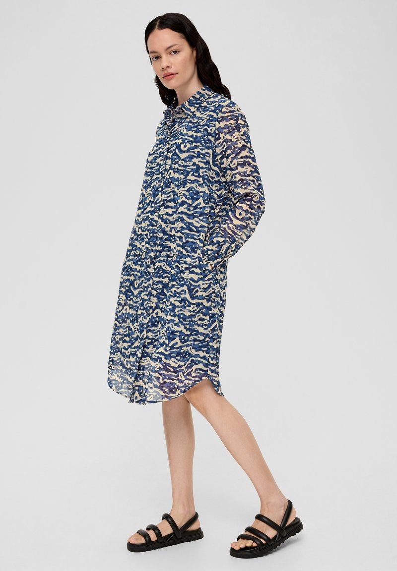 Robe chemise ample à motifs bleus et crème, à manches longues, avec un boutonnage sur le devant et des poches latérales. Portée avec des sandales noires à brides.