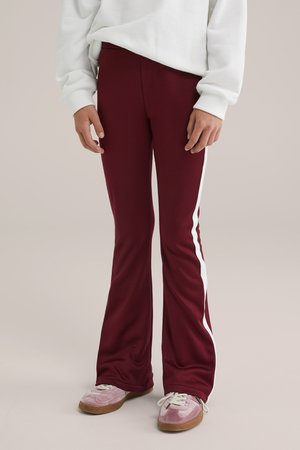 Pantaloni svasati bordeaux con strisce laterali bianche, realizzati in un tessuto morbido, dotati di una vita elastica e una texture liscia. Abbinati a sneakers rosa.