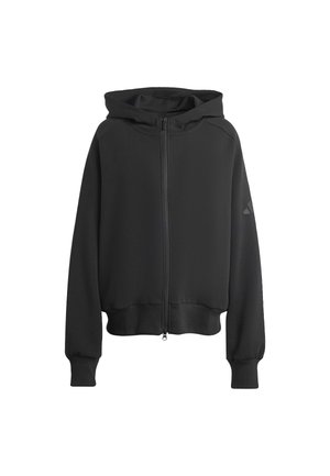 Junge Frau trägt eine schwarze Kapuzenjacke mit Reißverschluss und passende schwarze plissierte Shorts, steht vor einem schlichten hellen Hintergrund.