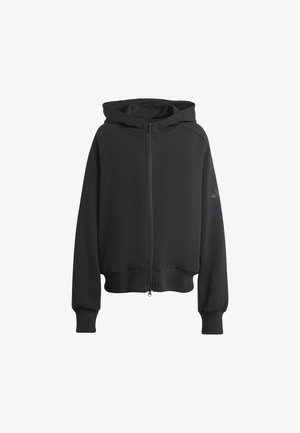 Junge Frau trägt eine schwarze Kapuzenjacke mit Reißverschluss und passende schwarze plissierte Shorts, steht vor einem schlichten hellen Hintergrund.