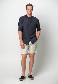Camicia azzurro navy in lino con colletto alla coreana, pantaloni corti crema e scarpe slip-on scure con suola in iuta. Design leggero e casual.