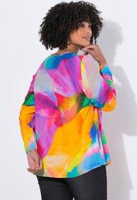 Vrouw met krullend haar die een kleurrijke, lange mouwen abstracte print top draagt, poseert met één hand bij haar gezicht en de andere hand achter haar rug.