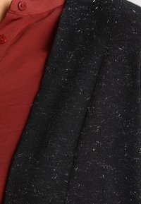 Tejido de punto negro con una superficie texturizada, con pequeñas fibras, mostrado junto a una camisa roja con botones.