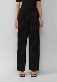Pantalon large noir plissé avec taille élastique, texture lisse et fines rayures verticales. Porté avec des sandales noires simples.