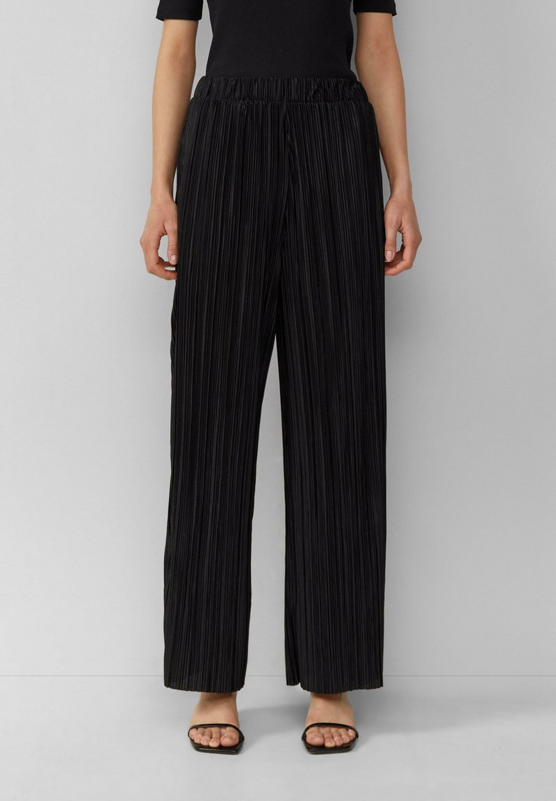 Pantalon large noir plissé avec taille élastique, texture lisse et fines rayures verticales. Porté avec des sandales noires simples.