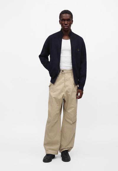 Homme portant une veste noire zippée par-dessus un débardeur blanc, un pantalon beige à jambes larges et des chaussures noires, debout devant un fond uni.