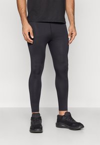 Férfi fekete sportos leggingsben és fekete futócipőben áll egy egyszerű világos háttér előtt.