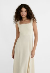 Beige linen dress with a square neckline and ruffled straps. It has a fitted bodice and a flowing skirt, featuring a side slit.