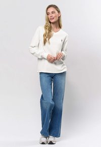 Vit långärmad V-ringad topp med ribbade mudd, kombinerad med ljusblå vida jeans och grå sportskor, mot en ljus bakgrund.