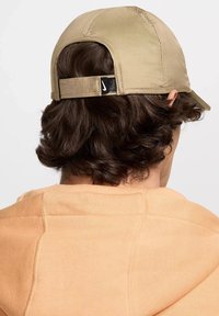 Cappellino da baseball beige con visiera curva, cinturino in tessuto regolabile e piccolo logo sul retro; visto da dietro con capelli ondulati visibili.