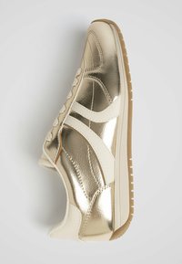 Metalen gouden sneaker met witte accenten, voorzien van een vetersluiting aan de voorkant en gestructureerde panelen, geplaatst op een lichtbruine rubberen zool.