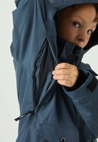 Giacca impermeabile blu navy con cerniera, tessuto strutturato, cappuccio regolabile e dettagli in velcro. Primo piano di una mano che si avvicina alla cerniera.