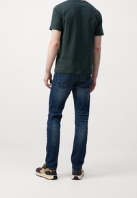 Man staat met zijn rug naar de camera, gekleed in een donkergroen T-shirt met korte mouwen, blauwe spijkerbroek en beige en zwarte sneakers, op een witte achtergrond.