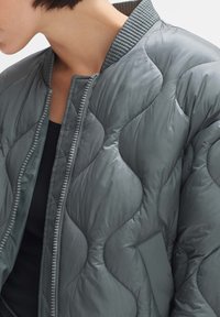 Gepaddede bomberjack in donkergrijs, met ribbelkraag, ritssluiting en een gladde, glanzende textuur met subtiele golvenpatroon details.