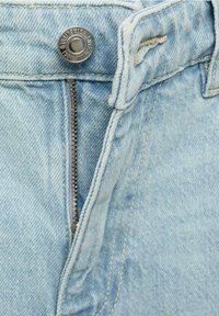 Gros plan sur un jean en denim bleu clair montrant une fermeture éclair métallique partiellement ouverte et un bouton argenté à la taille.