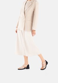 Sandalia con tiras que presenta una combinación de cuero nude y negro, con una suela robusta, un detalle de hebilla y un diseño de punta abierta.