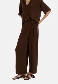Brązowa dzianinowa koszula z kołnierzykiem w kształcie wycięcia i zapięciem na guziki, w połączeniu z brązowymi spodniami w stylu owijanym oraz wzorzystymi sandałami z pomponami.