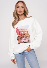 Biała bluza z nadrukiem przedstawiającym Koloseum oraz różowy samochód otoczony żywymi kwiatami, o luźnym kroju i ściągaczach przy mankietach.