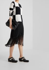 Svetr s černobílým kostkovaným vzorem, černá sukně se třásněmi, černá kabelka na zápěstí a masivní černé boty na podpatku s metalickým akcentem.