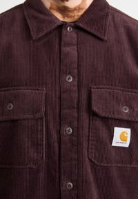 Mørk burgunder kordrowskjorte med spidse kraver, knaplukning fortil, to brystlommer og et firkantet logo-patch med et orange emblem.