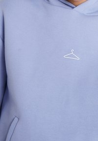 Sudadera ligera de color lavanda hecha de material de algodón suave, con un logo de perchero blanco bordado en el pecho y un bolsillo tipo canguro.