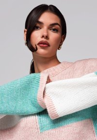 Nainen, jolla on tumma hius ja kultaiset rengaskorvakorut, pukeutuneena vaaleanpunaiseen, vihreään ja valkoiseen neulepaitaan, yksinkertaista vaaleaa taustaa vasten.