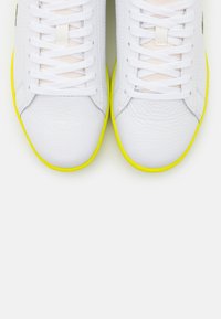 White sneakers made of textured leather, featuring a bright yellow rubber sole, flat white laces, and a minimalistic design.