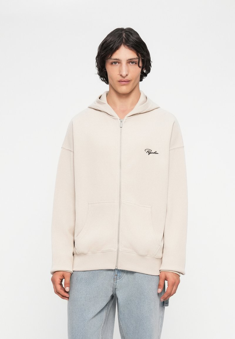 Beige mikina na zip s kapucí z ribbed látky, se přední kapsou a černě vyšitým logem na prsou. Nositelná s světle modrými džíny.