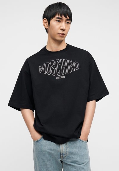 Μαύρο βαμβακερό μπλουζάκι με το λογότυπο "MOSCHINO" σε λευκό περίγραμμα και την ένδειξη "1983" κάτω από αυτό, άνετη εφαρμογή, κοντά μανίκια, συνδυασμένο με τζιν παντελόνι.