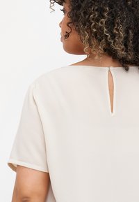 Beige blouse with a textured fabric, short sleeves, and a back keyhole opening secured by a button. Smooth drape and minimalistic design.