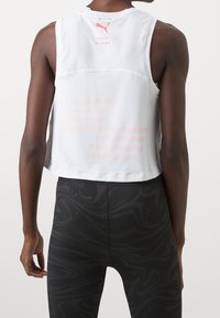 Valkoinen ilman hihoja urheiluyläosa, jossa on lyhennetty malli, pinkin värin gradientti raidat ja logo takana. Yhdistetty mustiin kuviollisiin legginsseihin.