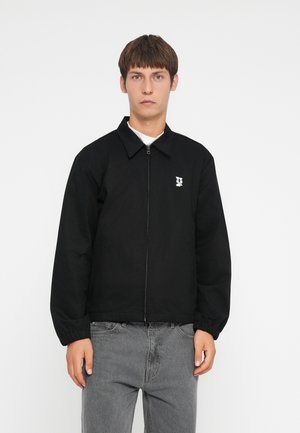 Black zippered jacket with a collared neckline, elastic cuffs, and a white logo on the left chest. Worn over a white shirt and gray jeans.