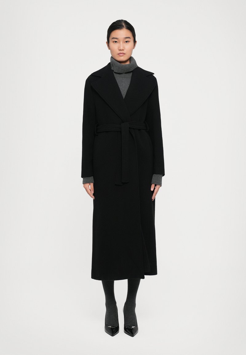 Woman wearing a long black belted coat over a gray turtleneck dress with matching socks and black pointed-toe heels, standing against white background.