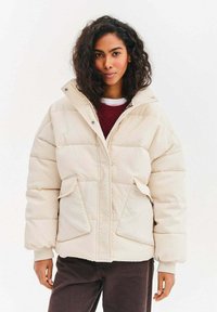 Prošívaná bunda krémové barvy s prošívanou texturou, vysokým límcem, předním zipem a dvěma velkými bočními kapsami. Nositelná přes vínový svetr.