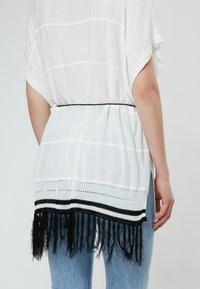 Witte gebreide top met een zwarte franjeszoom, met een losse pasvorm, wijde mouwen en zijsplitten, gedragen met lichtblauwe jeans.