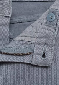 Tessuto in denim grigio con vista parziale di una zip, chiusura a bottone e dettagli cuciti. Texture liscia con sottili strisce.