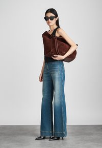 Καφέ σουέτ τσάντα tote με λεία υφή, μεγάλο μέγεθος και αντικρουόμενες δερμάτινες λεπτομέρειες. Το μοντέλο φοράει μαύρο αμάνικο τοπ και φαρδιά μπλε τζιν.