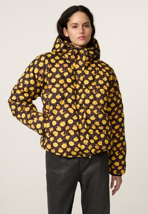 Vrouw die een gewatteerde jas met capuchon draagt met een geel bloemenpatroon en een zwarte broek, staand tegen een effen lichte achtergrond.