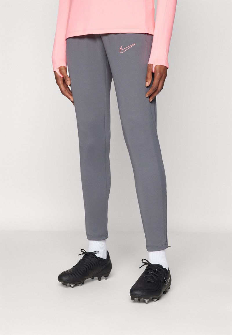Pantaloni sportivi gri cu o croială slim, având un logo Nike roz, asortați cu ghete de fotbal negre și șosete albe. Material moale cu o textură subtilă.