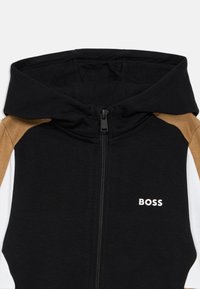 Casaco com capuz e fecho em preto, castanho e branco. Apresenta um material de malha flexível e um logótipo "BOSS" em branco na frente.