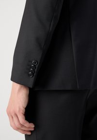 Maročni sakou z črno teksturo, ki je končan s štirimi črnimi gumbi in prilagojenim krojem. Roka položena na rokav.