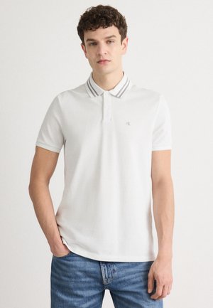 Mann in weißem Poloshirt mit gestreiftem Kragen und blauen Jeans, der mit einer Hand in der Tasche vor einem schlichten Hintergrund steht.