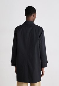 Abrigo negro de trench con un diseño estructurado, mangas largas y puños abotonados. Textura suave y forma de longitud media. Vista trasera sencilla.