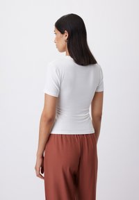Top aderente bianco a maniche corte con scollo rotondo, texture liscia e una leggera arricciatura in vita, abbinato a pantaloni marroni larghi.