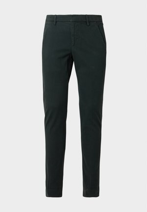 Homme debout portant un t-shirt à manches courtes vert olive, un pantalon slim vert foncé et des chaussures beiges à lacets sur fond blanc.