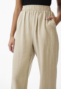 Woman wearing beige high-waisted wide-leg pants with elastic waistband, one hand in pocket and black top tucked in.