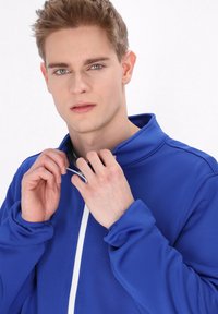 Giacca sportiva blu con colletto alto e zip bianca, dotata di maniche lunghe con polsini arricciati e un tessuto liscio e strutturato.